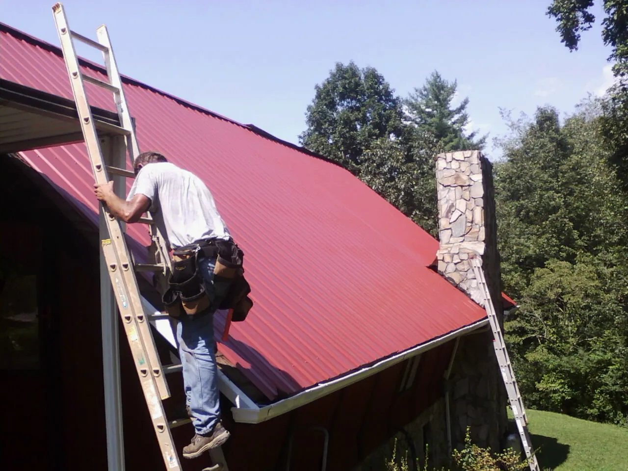 Roofer on a ladder beside a red metal roof and stone chimney — Soffit & Fascia Repair by Roofing Brothers in Villard